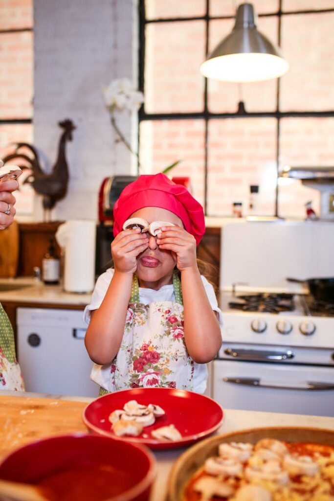 Young girl wearing a red chef hat and apron, playfully holding mushrooms in a cozy kitchen.